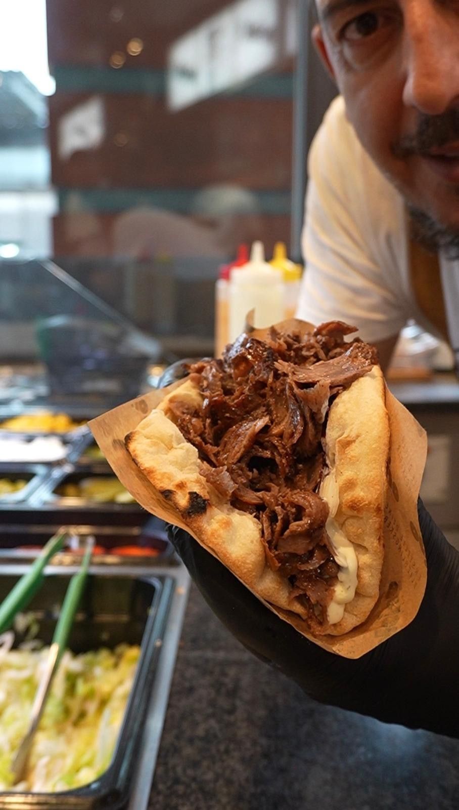 Döner im dicken, angeflämmten Brot mit Fleisch und weißer Sauce, von einer Hand im schwarzen Handschuh gehalten